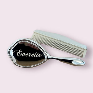 May include: A silver-coloured hairbrush and comb set. The brush has a black surface with the name "Everette" in white script. The brush is oval-shaped with a beaded border. The comb is white.