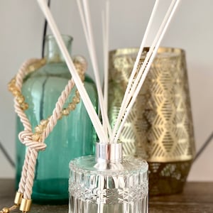 May include: A clear glass essential oil diffuser with white reeds, a silver collar, and a geometric pattern. A teal glass bottle and a gold geometric candle holder are in the background. A decorative rope with gold accents is draped over the teal bottle.