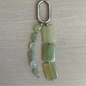 May include: A keychain featuring a silver-toned oval carabiner clip attached to two strands of light green stone beads. One strand consists of small oval-shaped beads, while the other strand features three larger rectangular-shaped stone pieces.