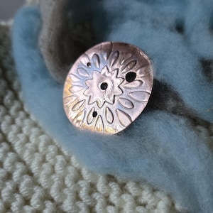 May include: A round copper-toned button with a stamped floral pattern and three holes, resting on a textured cream-colored knitted fabric layered with soft, light blue and grey felted wool fibers.