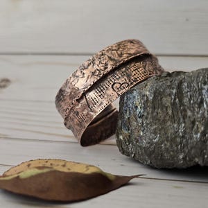 May include: Copper cuff bracelet with etched floral and heart designs. The bracelet is resting on a rock and a wooden surface. A dried leaf is in the foreground. The cuff is a warm, reddish-brown color.