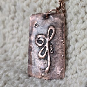May include: Copper-colored pendant necklace with a rectangular tag featuring a raised, stylized floral design. The pendant hangs from a matching copper chain. The background is a textured, off-white fabric.