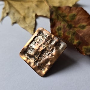 May include: A square-shaped metal ring with a hammered, textured surface featuring a warm copper-toned finish. The metal has a grid-like pattern of indentations across the top face, creating a rugged, uneven appearance with dark and light highlights.