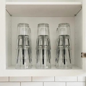 May include: Clear drinking glasses stacked inside a white cabinet. Each stack has a black, vertical support. The glasses are transparent, allowing visibility of the cabinet's interior. The cabinet is white with a white tiled backsplash.