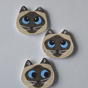 May include: Three hand-painted Siamese cat magnets. Each magnet features a tan face with dark brown markings, blue eyes, and white whiskers. The magnets are arranged on a white background.
