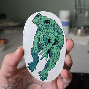 May include: An oval-shaped painting of a green and blue frog with detailed linework. The frog is depicted in a stylized, upright pose against a white background. The artwork has a handmade, artistic quality.