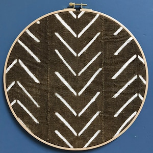 Puede incluir: Una tela marrón con un patrón de chevron blanco se estira sobre un aro de bordado de madera.