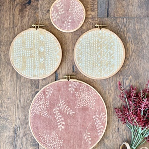 Pode incluir: Quatro aros de bordado redondos com padrões diferentes. Dois aros têm um padrão branco sobre um fundo bege, e dois aros têm um padrão branco sobre um fundo rosa.
