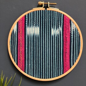May include: A round wooden embroidery hoop with a blue and white striped fabric with pink accents. The fabric has a woven texture.