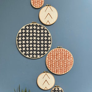 May include: Five round embroidery hoops with different geometric patterns in black, white, and brown on a blue wall. The hoops are hanging on the wall with a small plant in the bottom left corner.