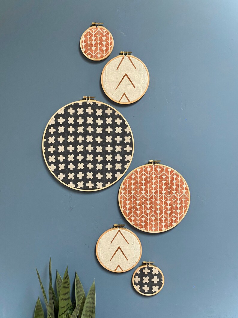 May include: Five round embroidery hoops with different geometric patterns in black, white, and brown on a blue wall. The hoops are hanging on the wall with a small plant in the bottom left corner.