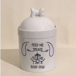 May include: White ceramic dog treat jar with a bone-shaped lid. The jar has a gray dog face design with the text "Feed me treats & tell me I am a good dog!"