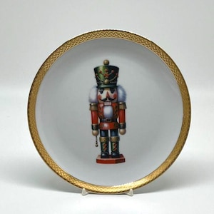 May include: A white decorative plate with a gold patterned rim. The plate features a colorful illustration of a nutcracker soldier in a red coat, black boots, and a green hat. The plate is displayed on a white stand.