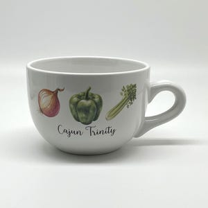 May include: White ceramic mug with a handle, featuring illustrations of an onion, green bell pepper, and celery. The words "Cajun Trinity" are written below the vegetables. The mug is designed for serving soups or beverages.