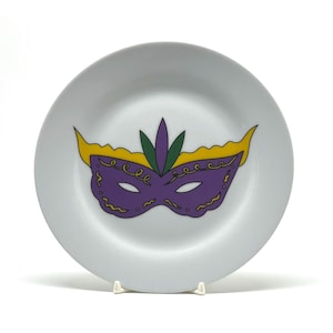 May include: A white ceramic plate featuring a purple, yellow, and green Mardi Gras mask design. The mask has a decorative swirl pattern and is centered on the plate. The plate is round and has a simple, elegant design.