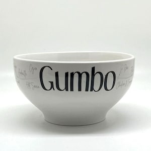 May include: White ceramic bowl with the word "Gumbo" printed in black. The bowl has a wide rim and a rounded bottom. The word "Gumbo" is centered on the bowl.