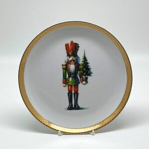 May include: A white ceramic plate with a gold rim featuring a detailed illustration of a nutcracker soldier holding a Christmas tree. The nutcracker is dressed in a red hat, green jacket, and red pants. The plate is supported by a white stand.