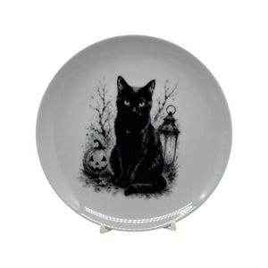 May include: A round light gray ceramic plate featuring a detailed black ink-style illustration of a sitting black cat, a carved jack-o'-lantern pumpkin, a vintage-style lantern, and bare tree branches centered on the surface of the plate.