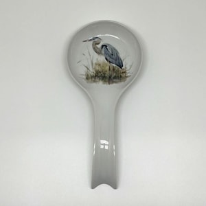 May include: A ceramic spoon rest with a gray handle and a round bowl. The bowl features a detailed illustration of a blue heron standing in a marshy area with tall grass. The spoon rest is white with a glossy finish.