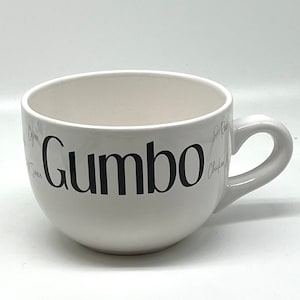 May include: White ceramic soup bowl with a black "Gumbo" inscription. The bowl has a handle and is designed for serving soup or stew.