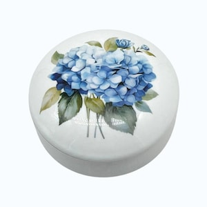 May include: A round white ceramic trinket box featuring a detailed illustration of blue hydrangea flowers with green leaves on the lid. The container has a smooth, glossy finish and a circular shape, providing a compact storage space for small items.