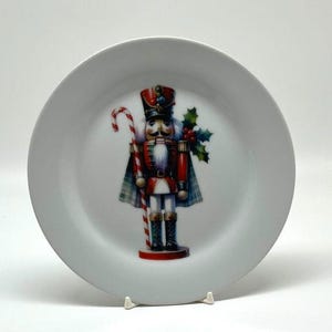 May include: White ceramic plate featuring a colorful illustration of a nutcracker soldier. The nutcracker is dressed in a red and black uniform, holding a candy cane. Holly and berries are also depicted on the plate, suggesting a holiday theme.