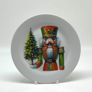 May include: A white ceramic plate featuring a colorful illustration of a Christmas nutcracker soldier holding a decorated Christmas tree. The nutcracker is dressed in red, green, and gold. The plate is round and has a simple, elegant design.