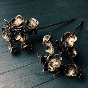 May include: Two metal flower arrangements against a dark blue backdrop. The flowers are crafted from metal and have a gold finish. The flowers are clustered together and have a delicate, feminine appearance.