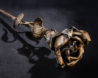 May include: A single golden rose made of metal with a long stem. The rose is lying on a dark gray surface.