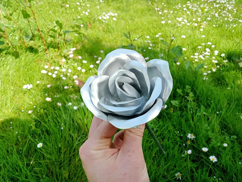 Pode incluir: Uma grande rosa de metal prateado com p&eacute;talas detalhadas. A rosa &eacute; segurada contra um fundo de grama verde e pequenas flores brancas. A haste &eacute; de cor verde escuro.