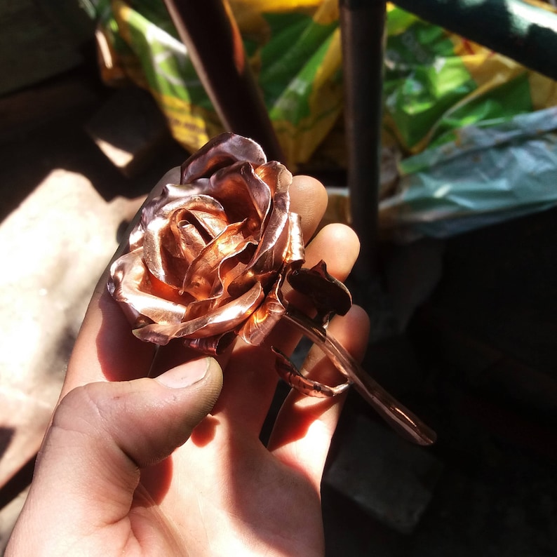 May include: A copper rose sculpture with a detailed, realistic design. The rose is crafted from hammered copper, giving it a textured, metallic finish.