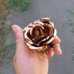 May include: A copper rose sculpture with a detailed, realistic design. The rose is crafted with multiple layers of petals, creating a three-dimensional effect.