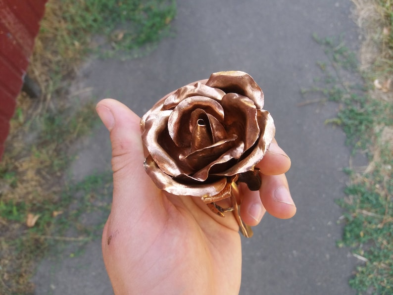 May include: A copper rose sculpture with a detailed, realistic design. The rose is crafted with multiple layers of petals, creating a three-dimensional effect.