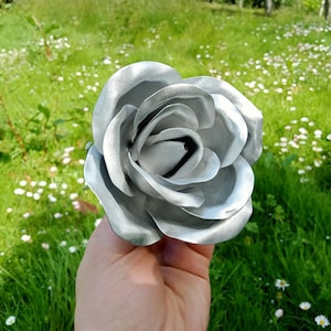 Pode incluir: Uma grande rosa prateada com m&uacute;ltiplas p&eacute;talas. A flor artificial &eacute; segurada em uma m&atilde;o contra um fundo de grama verde e pequenas flores brancas. As p&eacute;talas da rosa t&ecirc;m um leve brilho.