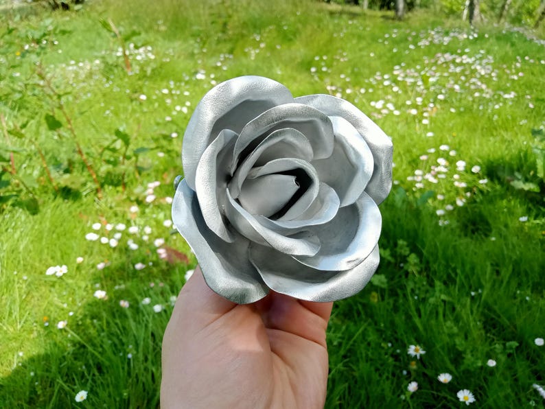 Pode incluir: Uma grande rosa prateada com m&uacute;ltiplas p&eacute;talas. A flor artificial &eacute; segurada em uma m&atilde;o contra um fundo de grama verde e pequenas flores brancas. As p&eacute;talas da rosa t&ecirc;m um leve brilho.
