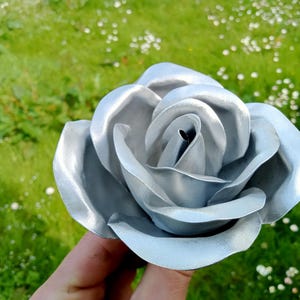 Pode incluir: Uma rosa de metal prateado com detalhes intrincados nas p&eacute;talas. A rosa &eacute; segurada em frente a um fundo de relva verde e pequenas flores brancas. O acabamento met&aacute;lico confere &agrave; flor um aspeto moderno.