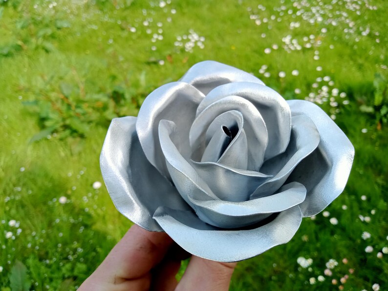 Pode incluir: Uma rosa de metal prateado com detalhes intrincados nas p&eacute;talas. A rosa &eacute; segurada em frente a um fundo de relva verde e pequenas flores brancas. O acabamento met&aacute;lico confere &agrave; flor um aspeto moderno.