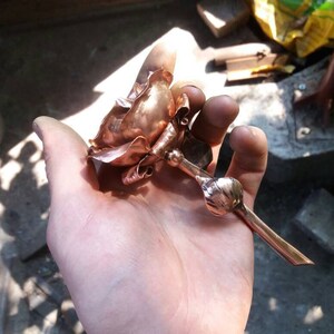 May include: A copper rose sculpture with a long stem, held in a hand. The rose is made of thin, hammered copper and has a realistic shape.