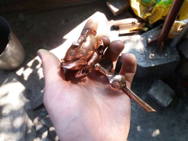 May include: A copper rose sculpture with a long stem, held in a hand. The rose is made of thin, hammered copper and has a realistic shape.