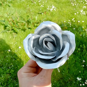Pode incluir: Uma grande rosa prateada met&aacute;lica com p&eacute;talas detalhadas. A flor artificial &eacute; segurada contra um fundo de grama verde e pequenas flores brancas, criando um contraste de texturas e cores. A rosa est&aacute; totalmente aberta.