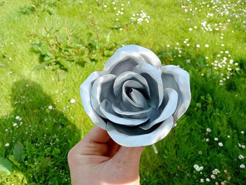 Pode incluir: Uma grande rosa prateada met&aacute;lica com p&eacute;talas detalhadas. A flor artificial &eacute; segurada contra um fundo de grama verde e pequenas flores brancas, criando um contraste de texturas e cores. A rosa est&aacute; totalmente aberta.