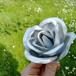 Pode incluir: Um close-up de uma rosa prateada com p&eacute;talas detalhadas. A flor artificial &eacute; segurada contra um fundo de grama verde e pequenas flores brancas. O brilho met&aacute;lico da rosa confere-lhe uma apar&ecirc;ncia &uacute;nica e elegante.