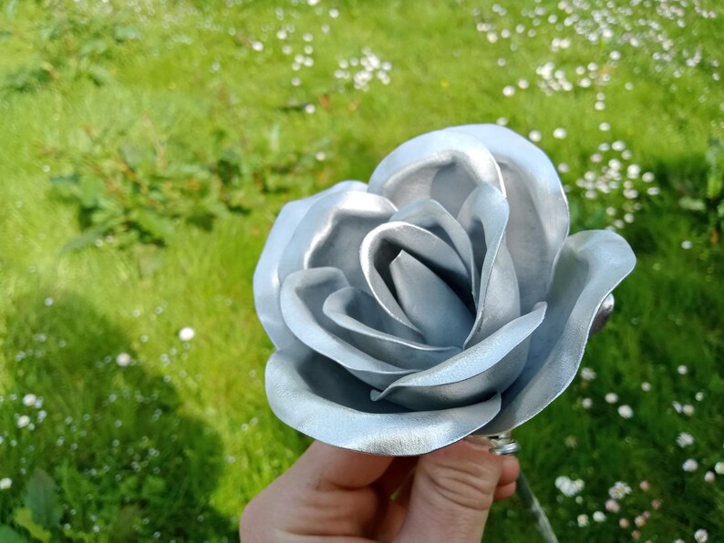 Pode incluir: Um close-up de uma rosa prateada com p&eacute;talas detalhadas. A flor artificial &eacute; segurada contra um fundo de grama verde e pequenas flores brancas. O brilho met&aacute;lico da rosa confere-lhe uma apar&ecirc;ncia &uacute;nica e elegante.
