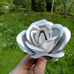 Pode incluir: Uma escultura de rosa em metal prateado com p&eacute;talas detalhadas. A flor artesanal &eacute; segurada contra um fundo de relva verde e &aacute;rvores. O brilho met&aacute;lico da escultura reflete a luz do sol, destacando o seu design intrincado.