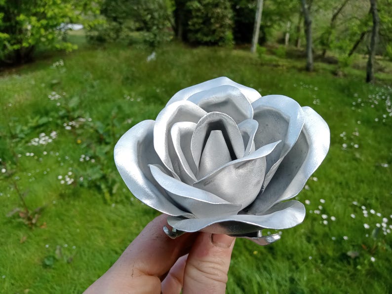 Pode incluir: Uma escultura de rosa em metal prateado com p&eacute;talas detalhadas. A flor artesanal &eacute; segurada contra um fundo de relva verde e &aacute;rvores. O brilho met&aacute;lico da escultura reflete a luz do sol, destacando o seu design intrincado.