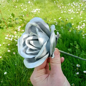 Pode incluir: Uma rosa de metal prateado com detalhes intrincados nas p&eacute;talas e um caule verde. A flor &eacute; colocada sobre um fundo de relva verde e pequenas flores brancas, criando um contraste natural.