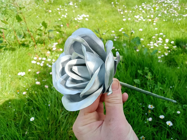 Pode incluir: Uma rosa de metal prateado com detalhes intrincados nas p&eacute;talas e um caule verde. A flor &eacute; colocada sobre um fundo de relva verde e pequenas flores brancas, criando um contraste natural.