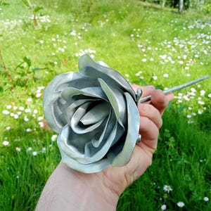 Pode incluir: Uma rosa de metal prateado com p&eacute;talas detalhadas, segurada contra um fundo de relva verde e pequenas flores brancas. A haste tamb&eacute;m &eacute; prateada. A rosa &eacute; um item decorativo feito &agrave; m&atilde;o.