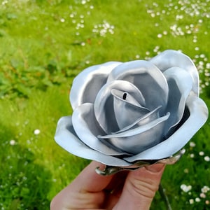 Pode incluir: Uma rosa de metal prateado com p&eacute;talas detalhadas. A rosa &eacute; segurada em frente a um fundo gramado verde. A rosa de metal &eacute; um item decorativo, possivelmente para um jardim ou decora&ccedil;&atilde;o de casa.