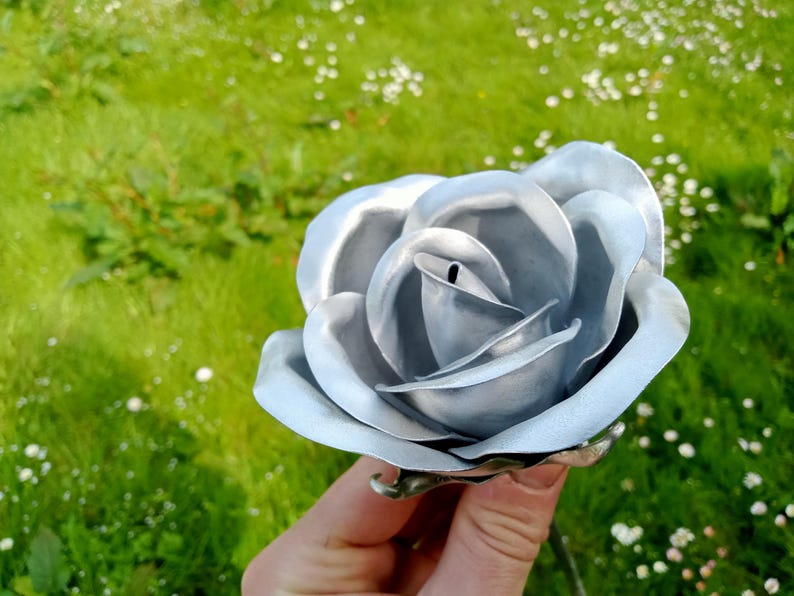 Pode incluir: Uma rosa de metal prateado com p&eacute;talas detalhadas. A rosa &eacute; segurada em frente a um fundo gramado verde. A rosa de metal &eacute; um item decorativo, possivelmente para um jardim ou decora&ccedil;&atilde;o de casa.